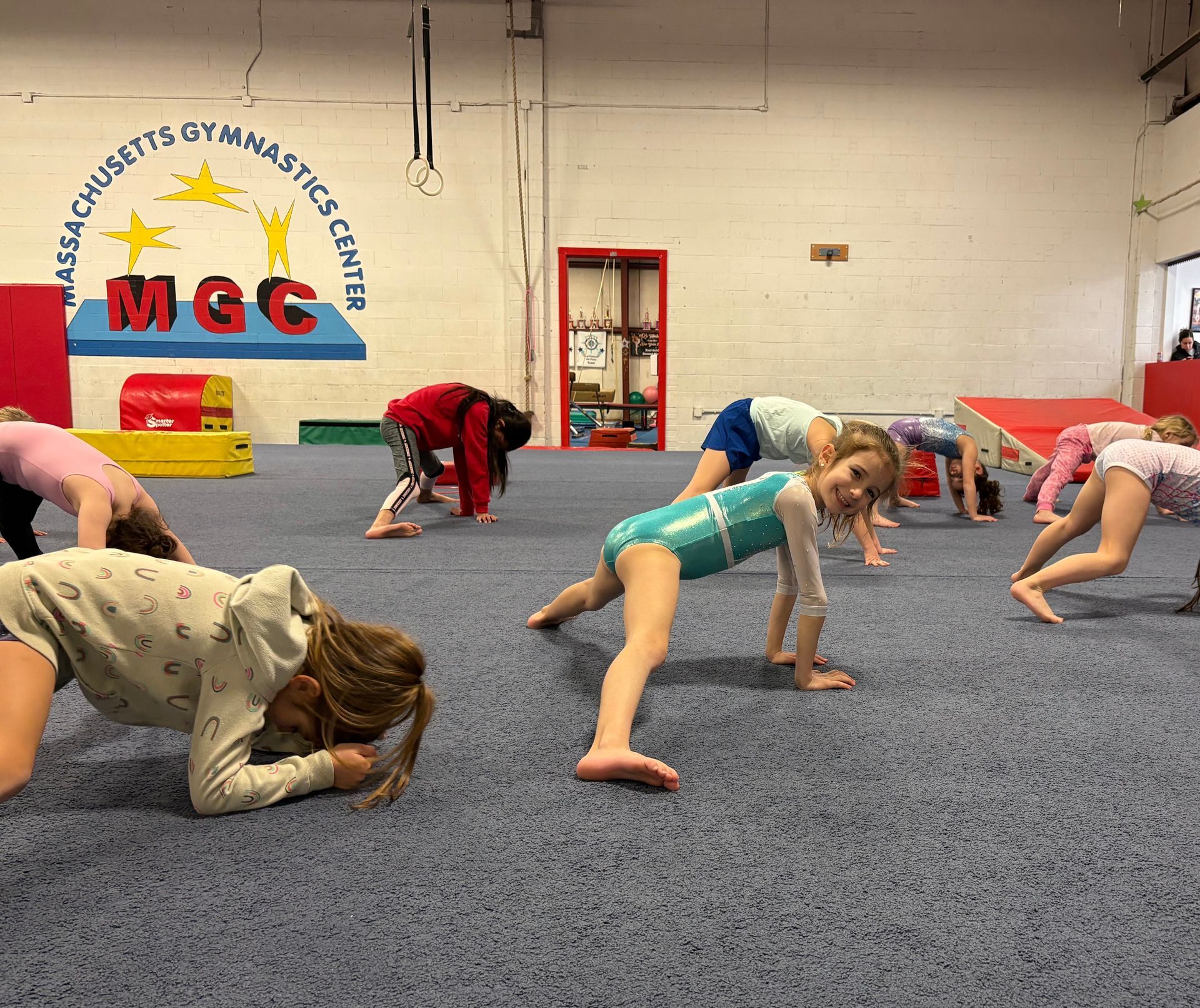 Children perform stretching exercises on the blue floor of a gymnastics center.