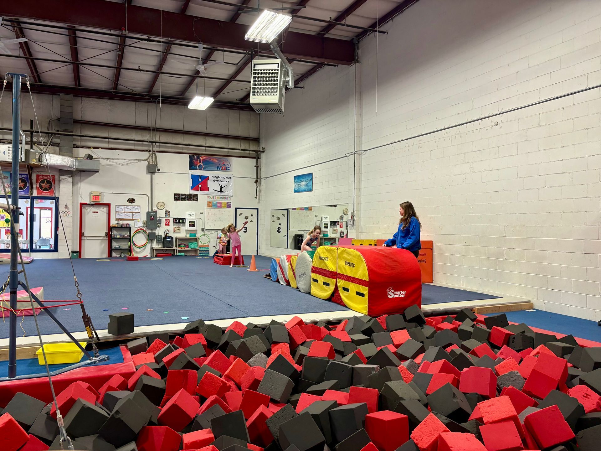A gym with a foam block pit in the foreground and a blue mat area with gymnastics equipment and people in the background.