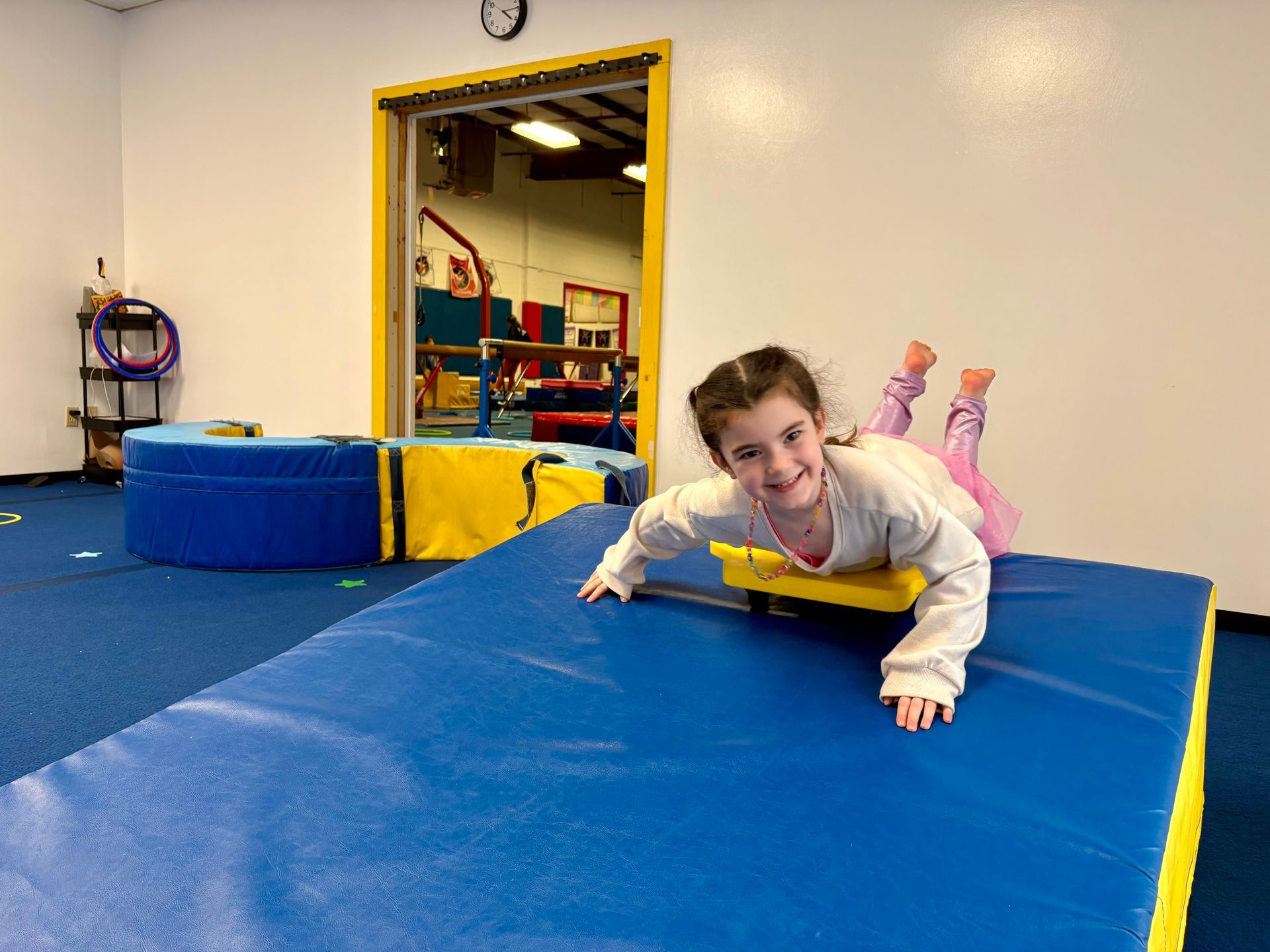 A child lies on their stomach on a yellow scooter atop a blue gym mat in an indoor gymnastics facility.