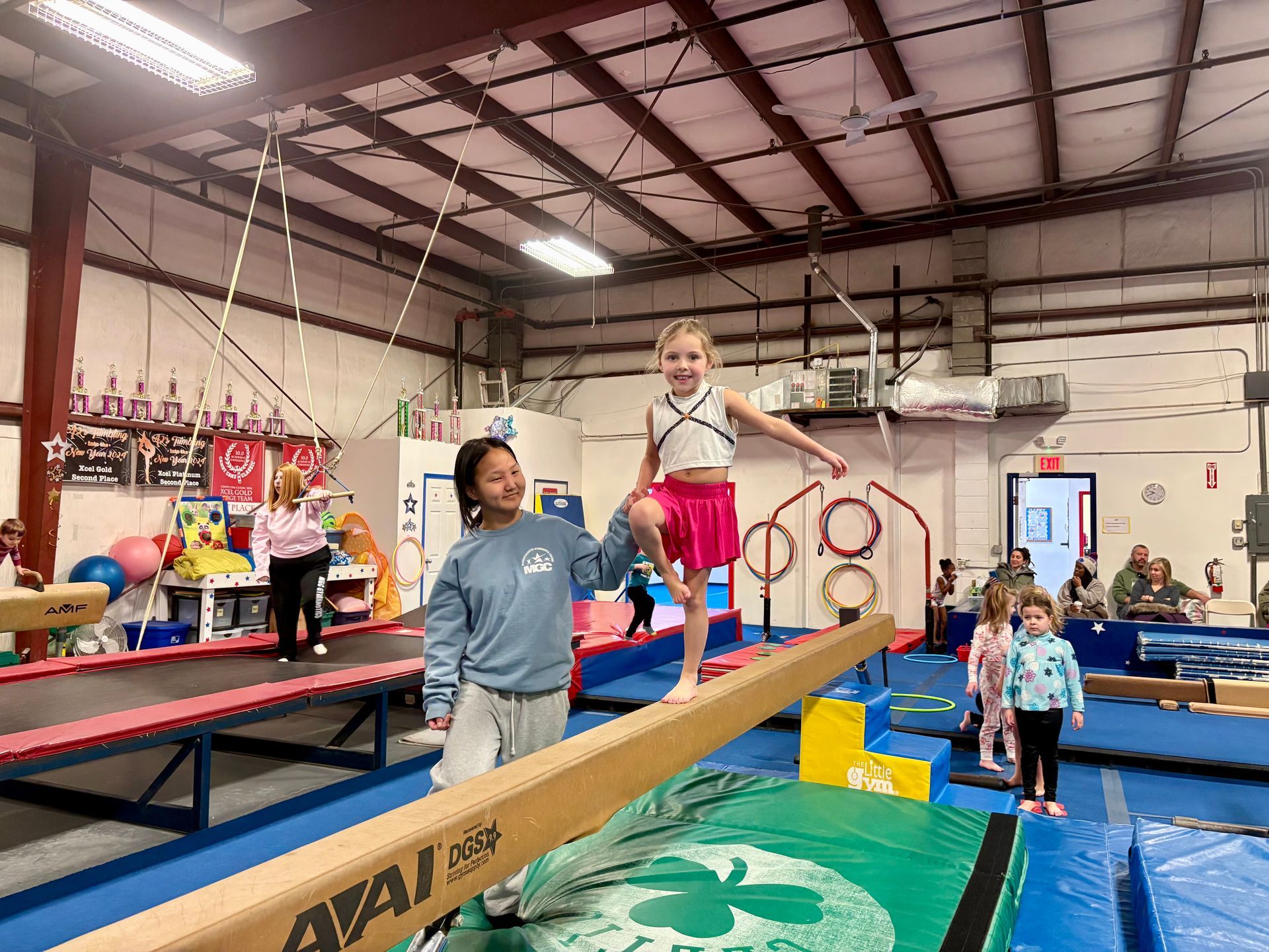 An instructor assists a student balancing on a gymnastics beam inside a gym.