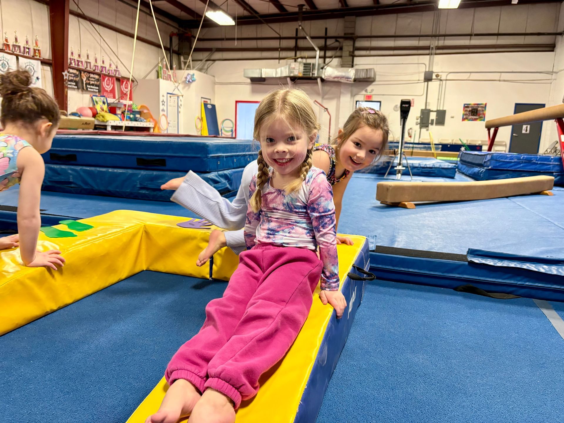 Three children sitting on yellow gymnastics mats in a training center, smiling and looking toward the camera.