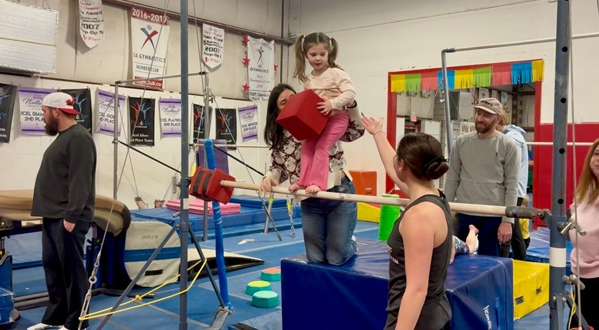 A child is helped across a balance beam in a gymnastics facility, with adults supervising nearby.