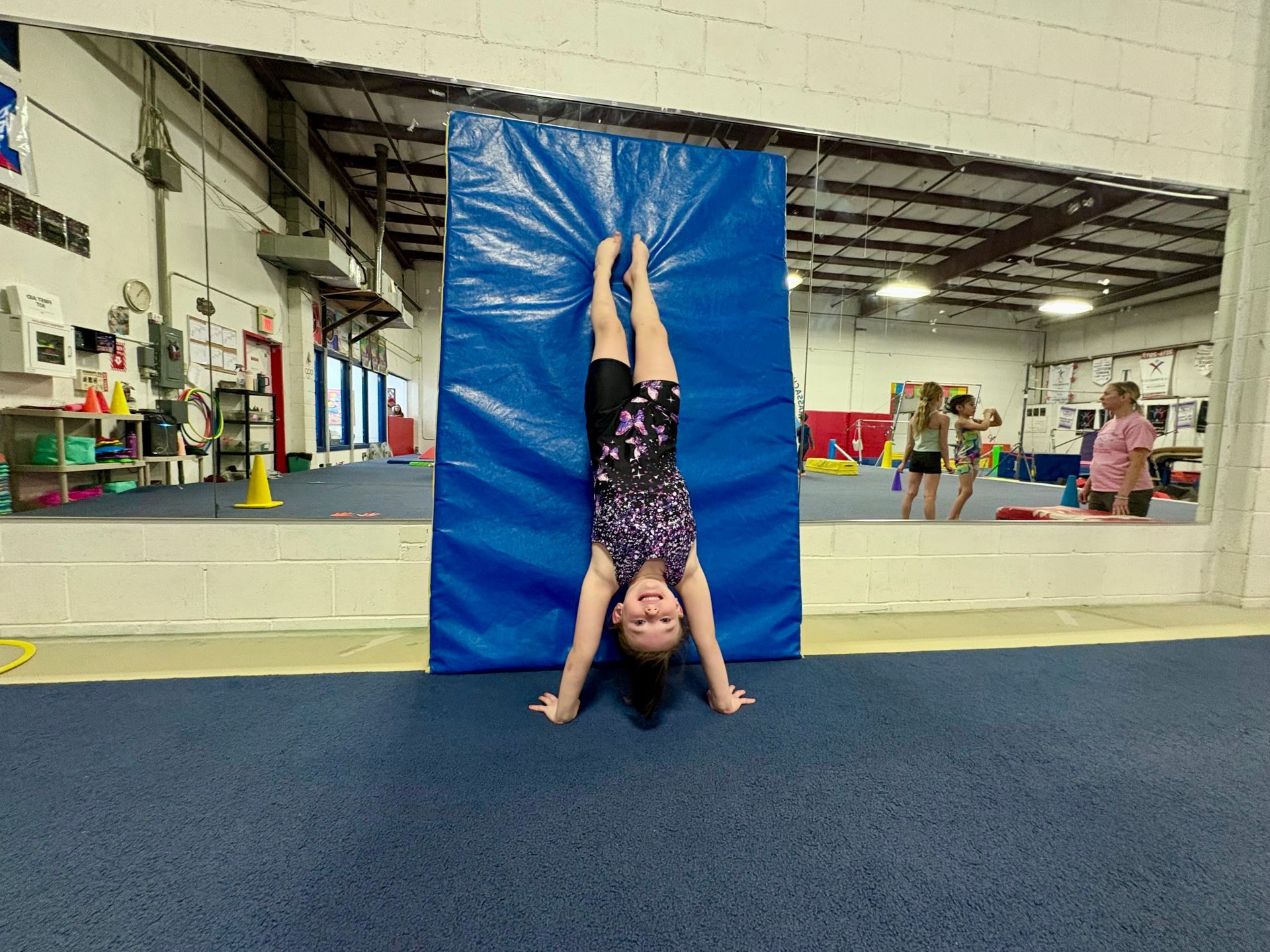 A person in a patterned leotard performs a handstand against a blue gym mat in an indoor gymnastics facility.