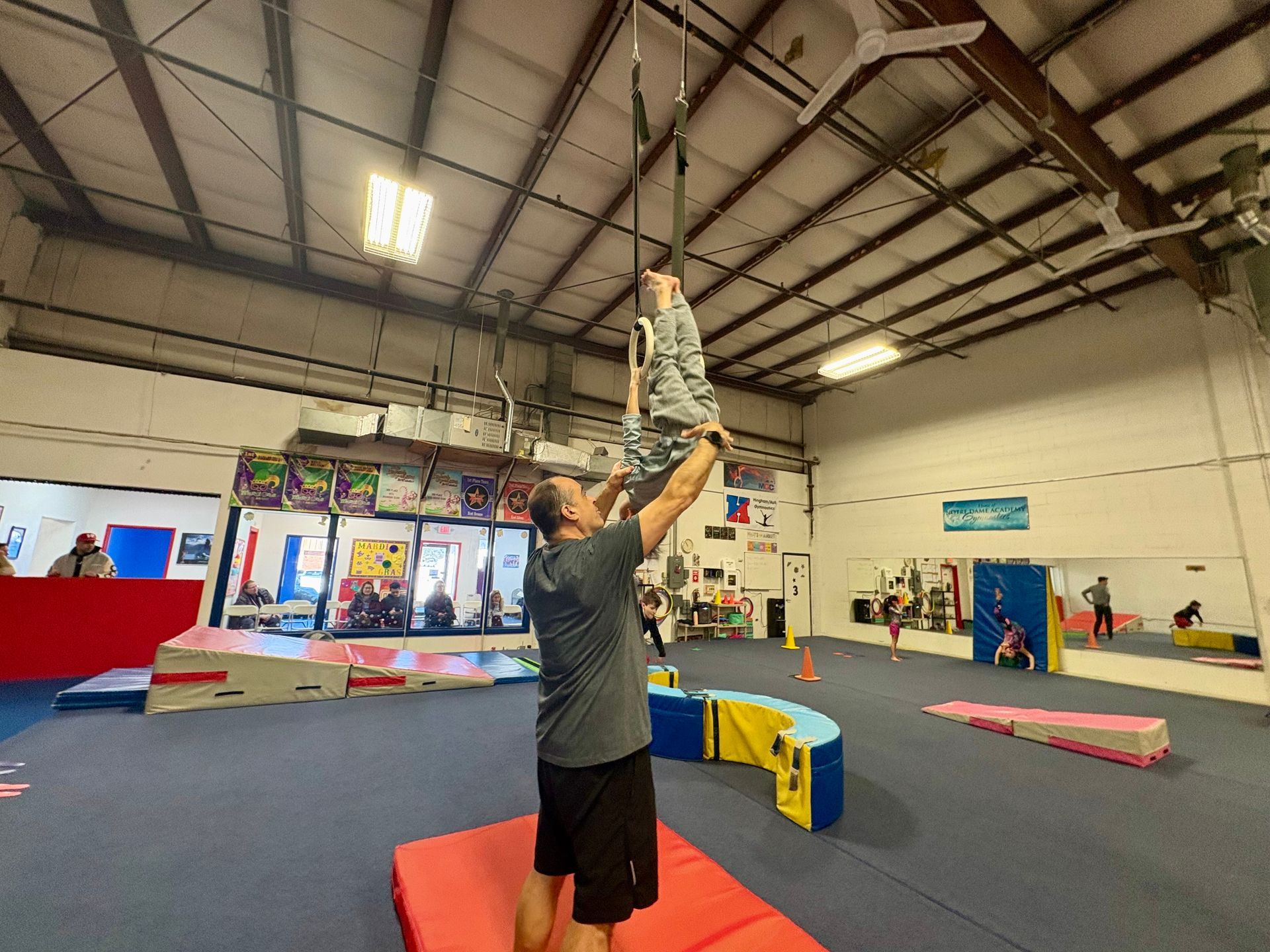 A man assists a person hanging from gymnastic rings in an indoor training facility.