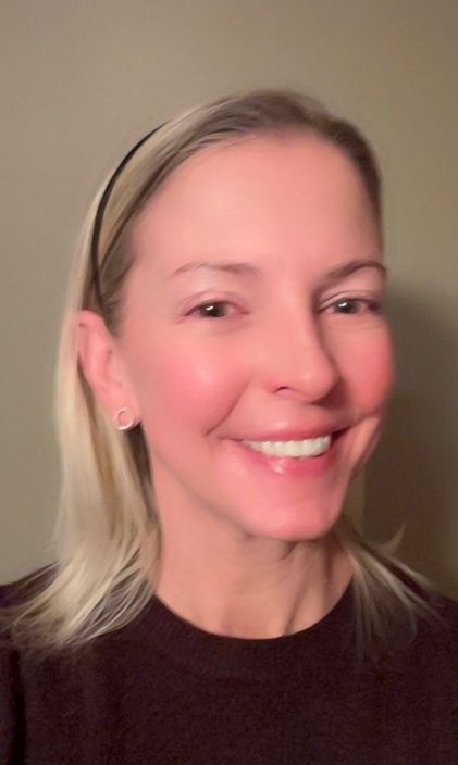 A person with blonde hair pulled back by a headband smiling at the camera, wearing a dark top against a plain wall.