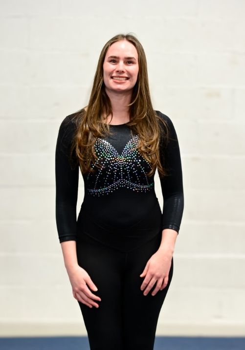 A smiling person wearing a black, long-sleeved leotard with a sequined pattern on the chest, standing against a white wall.