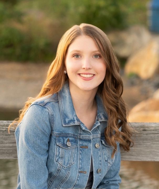 A young woman with long wavy brown hair smiles at the camera, wearing a blue denim jacket outdoors by a wooden railing.