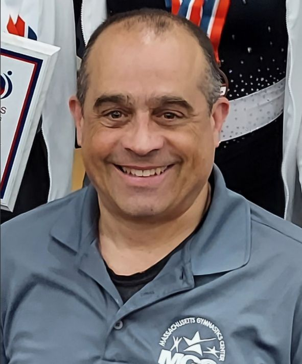 Smiling person wearing a gray polo shirt with a gymnastics center logo, standing in a gym setting.