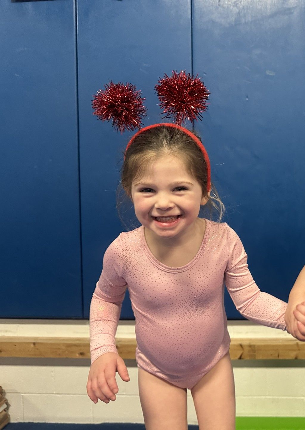 A smiling child in a light pink leotard wears red glittery pom-pom ears against a blue wall.