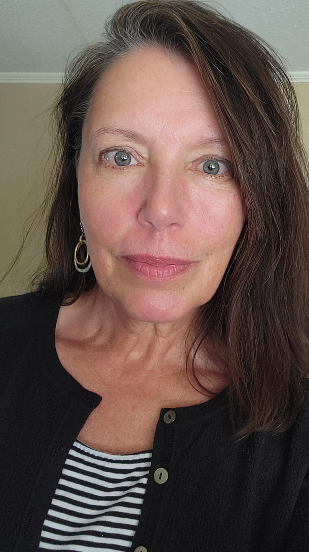 Woman with long brown hair, wearing a black cardigan over a striped shirt, and hoop earrings.
