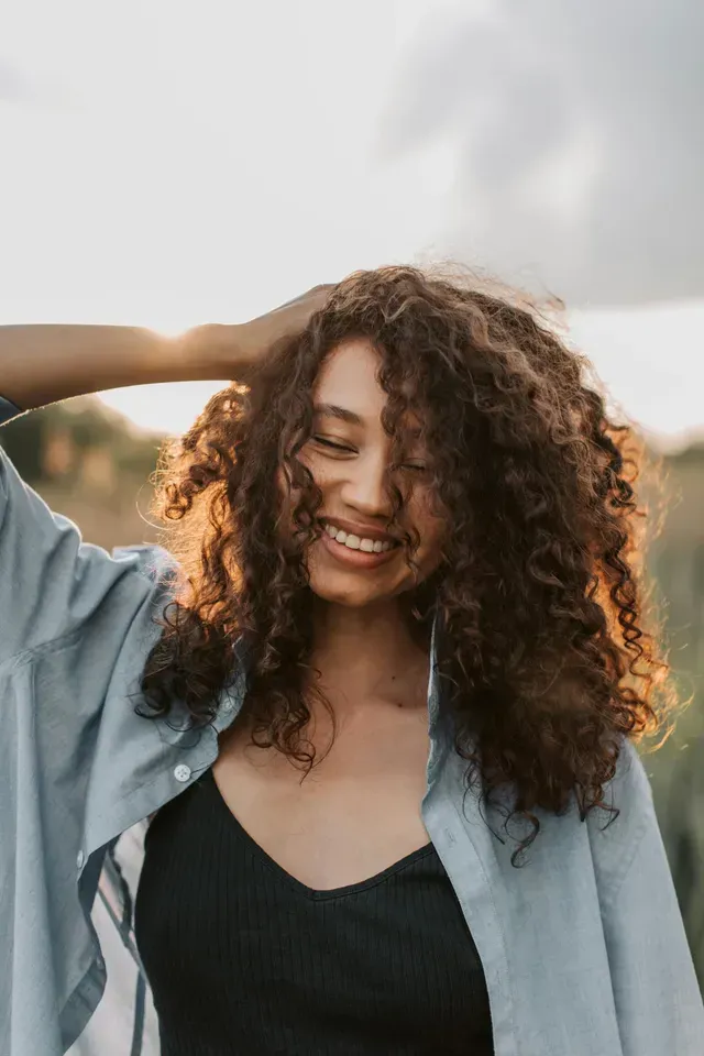 A woman with curly hair is smiling and touching her hair.
