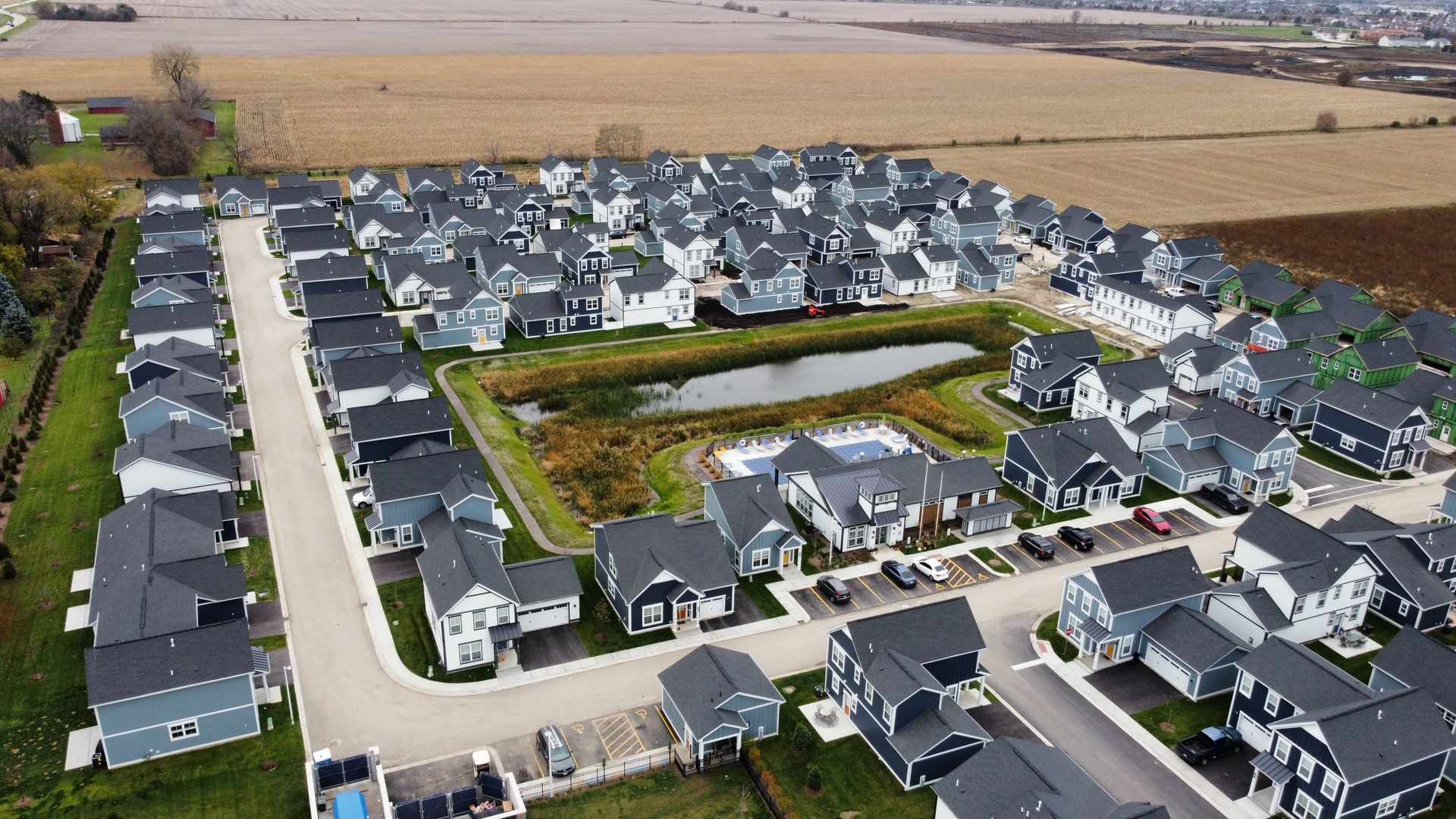 Aerial view of a suburban housing development with rows of gray-roofed homes and nearby farmland.