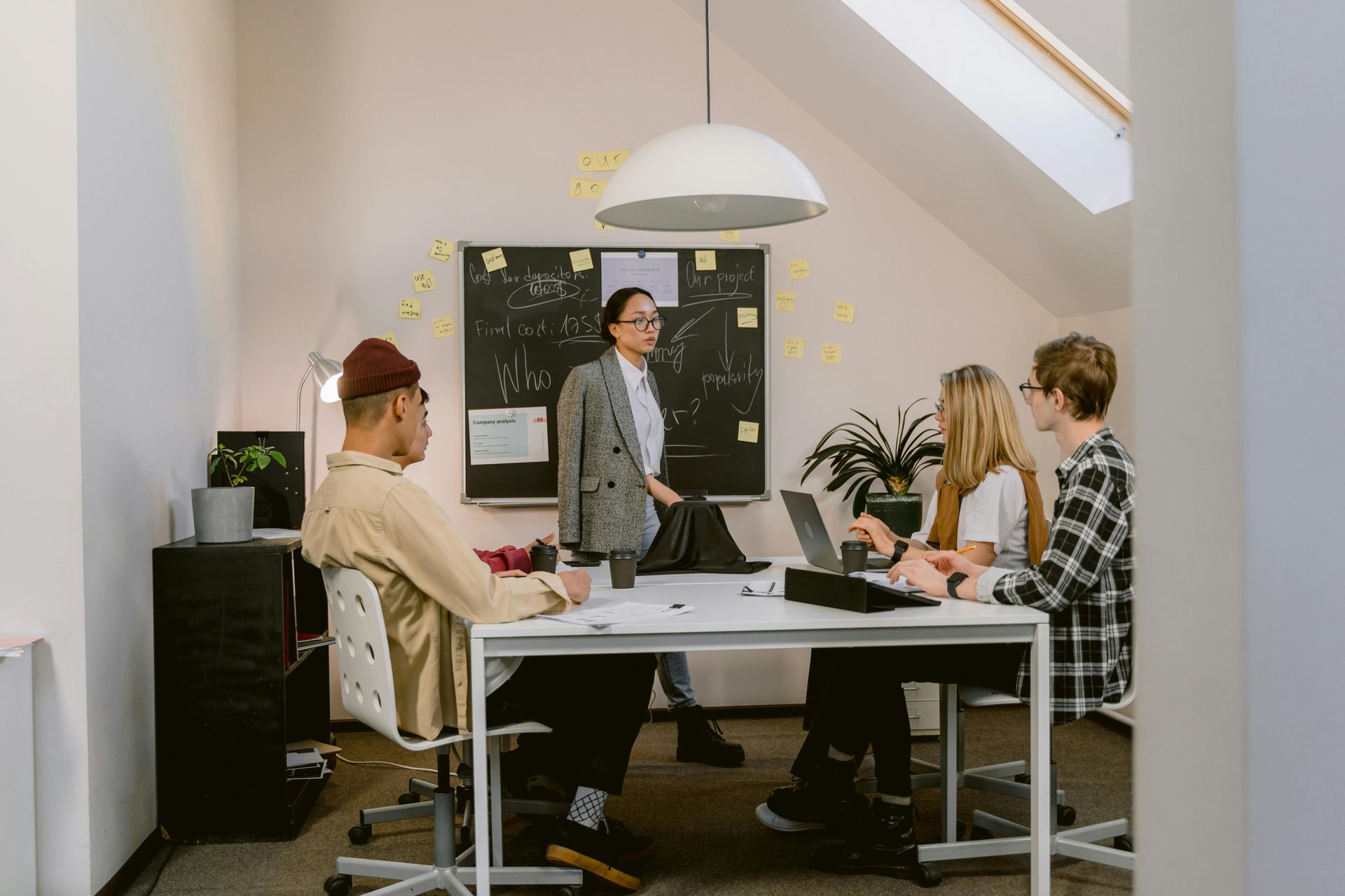 Cuatro personas en una reunión. Una mujer presenta en una pizarra, tres en una mesa con portátiles. Sala blanca.