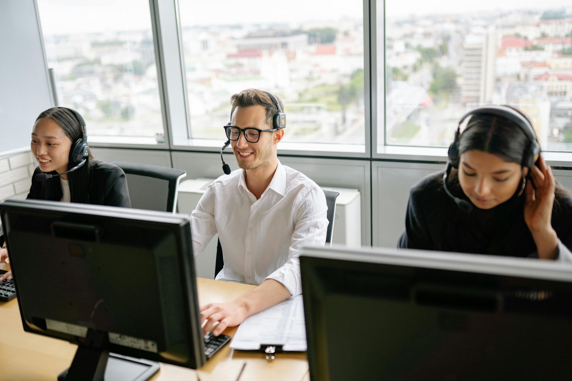Tres personas que usan auriculares trabajan con computadoras en una oficina con vista a la ciudad.