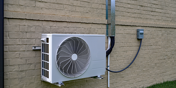 Outdoor wall-mounted air conditioner unit with visible fan and electrical conduit on a brick wall.