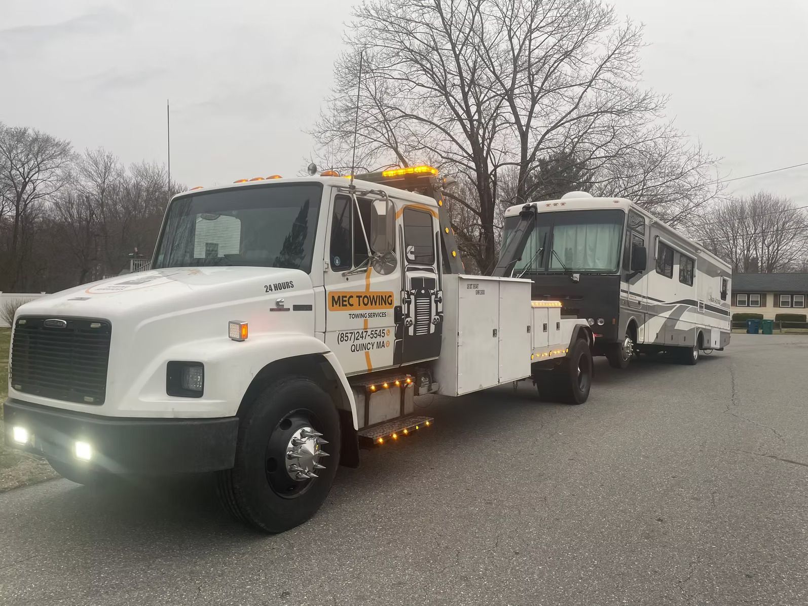 A white tow truck is towing a rv in a parking lot.