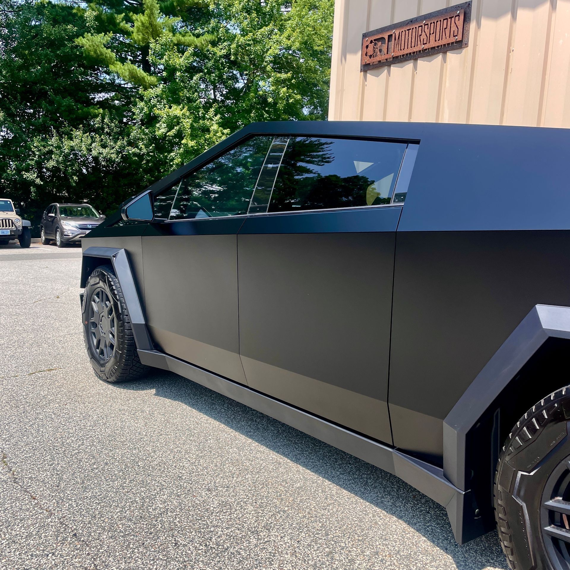 A matte black Tesla Cybertruck parked on a gravel lot near a beige building on a sunny day.