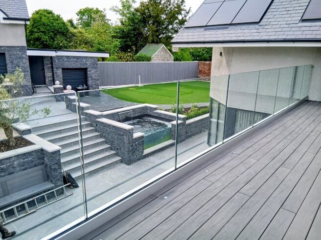 Modern deck with glass balustrade railing installed by Swansea Glass Balustrades. Hendrefoilan property overlooks a terraced backyard with steps, pool, and lawn.