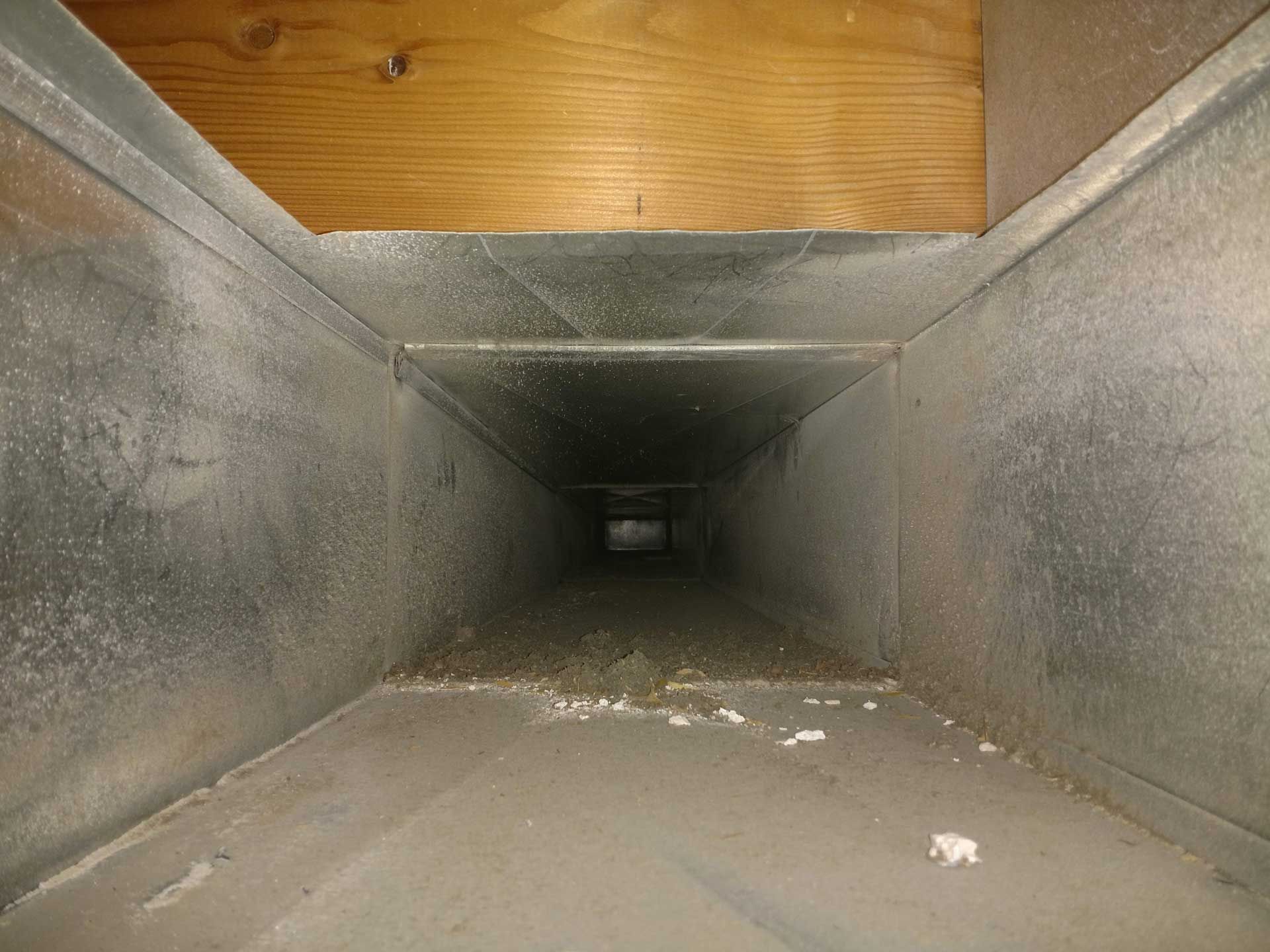 Looking down a dusty rectangular HVAC duct made of metal, wood above.