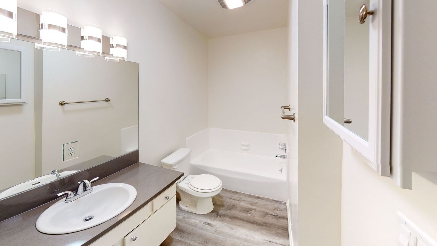 Bathroom with a white tub, toilet, sink, and mirror. Gray countertop and light-colored flooring.
