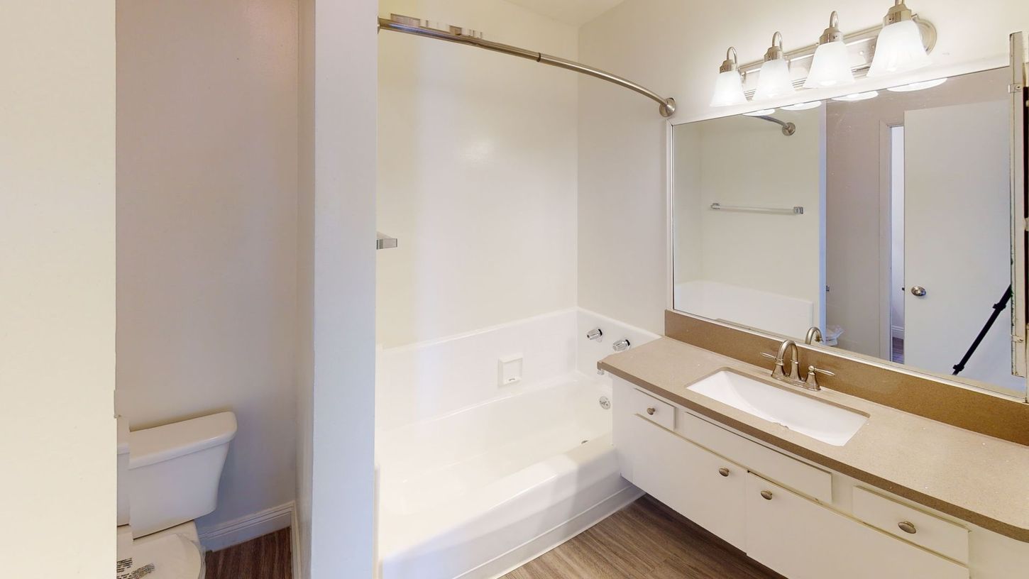 Bathroom with white tub, vanity, toilet, and a large mirror. Beige countertop and light wood-look floor.