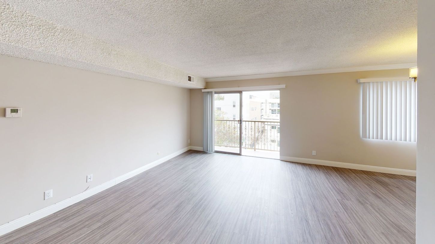 Empty living room with light-colored walls, wood-look flooring, and a sliding glass door leading to a balcony.