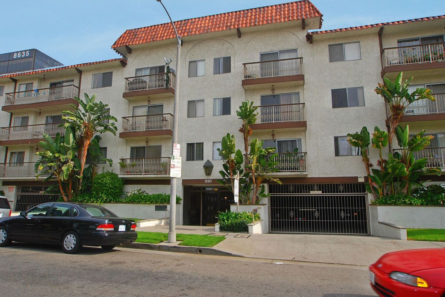 Four-story apartment building with balconies, brown trim, and a red tile roof. Cars parked in front.