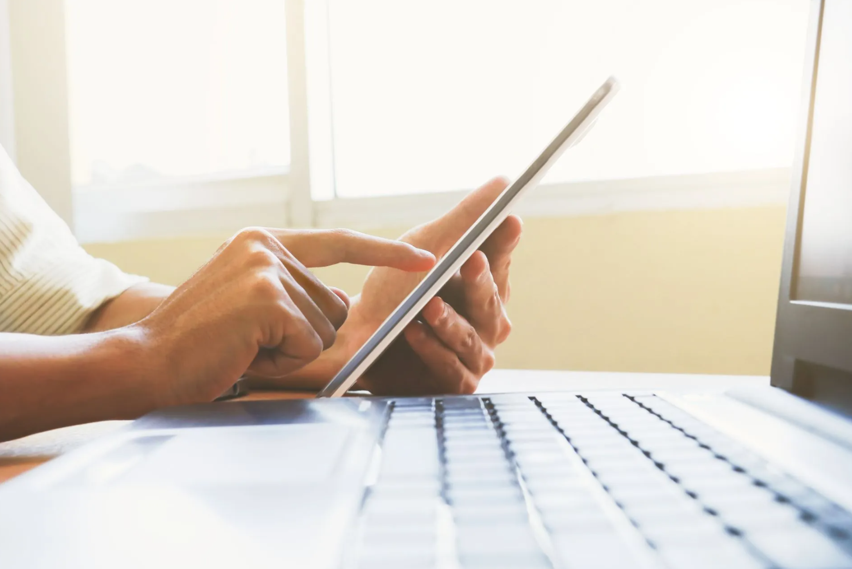 Person using a tablet and laptop near a window. Sunlight illuminates the scene.