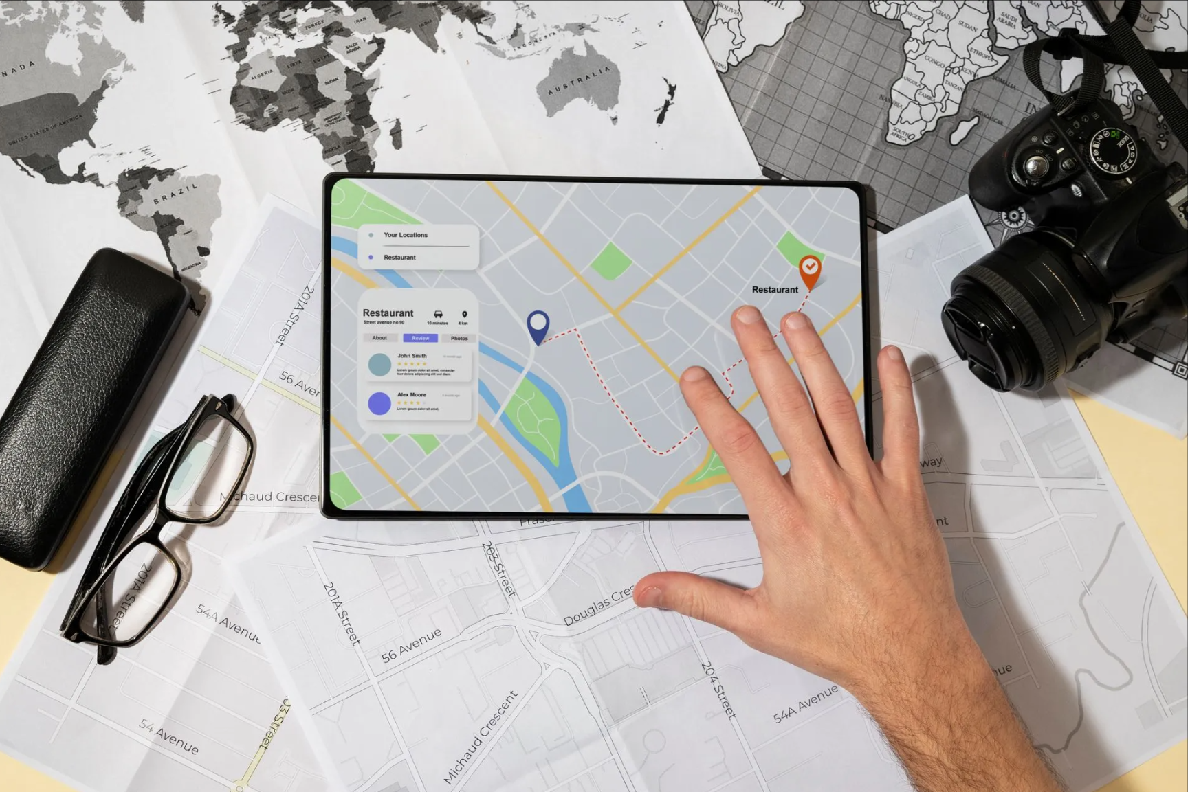 Person's hand pointing at map on a tablet. Nearby, a camera, glasses, map, and case are arranged on the table.