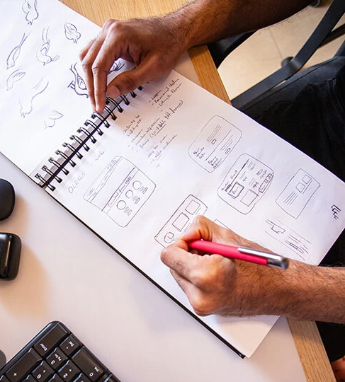 Person sketching UI design concepts in a spiral notebook with a pink pen.