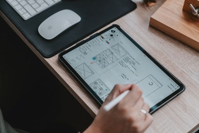 Person uses stylus to draw on tablet displaying a wireframe design on a wooden desk.