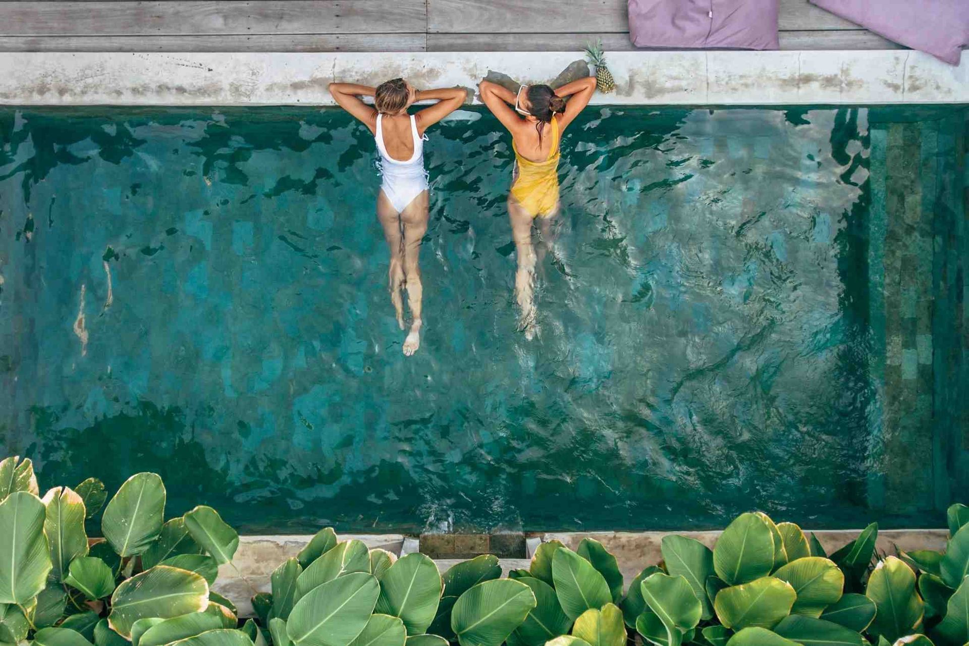 Two women in swimsuits relaxing in a turquoise pool, shot from above.