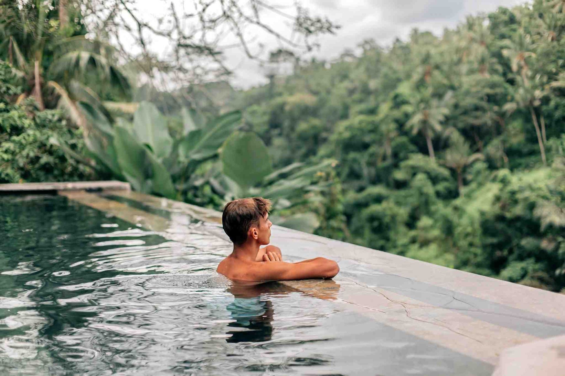 Man in an infinity pool, looking out at a lush green jungle landscape.