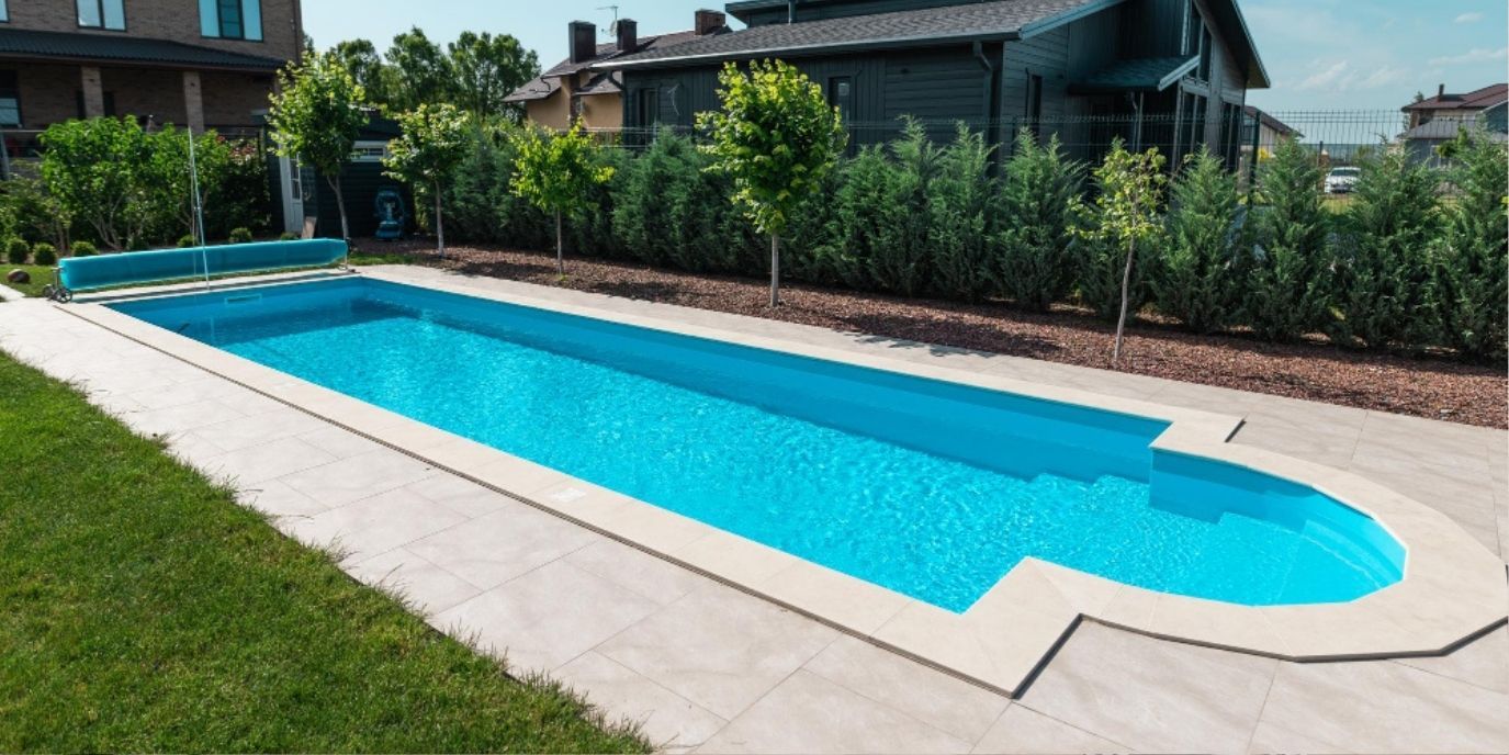 Modern rectangular swimming pool with bright blue water beside a white deck and green lawn, near homes