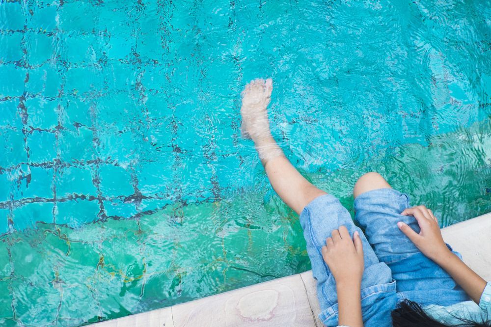 A person is sitting on the edge of a swimming pool with their legs in the water.