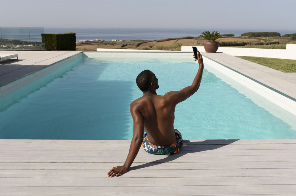 A man is sitting on the edge of a swimming pool taking a selfie with his phone.