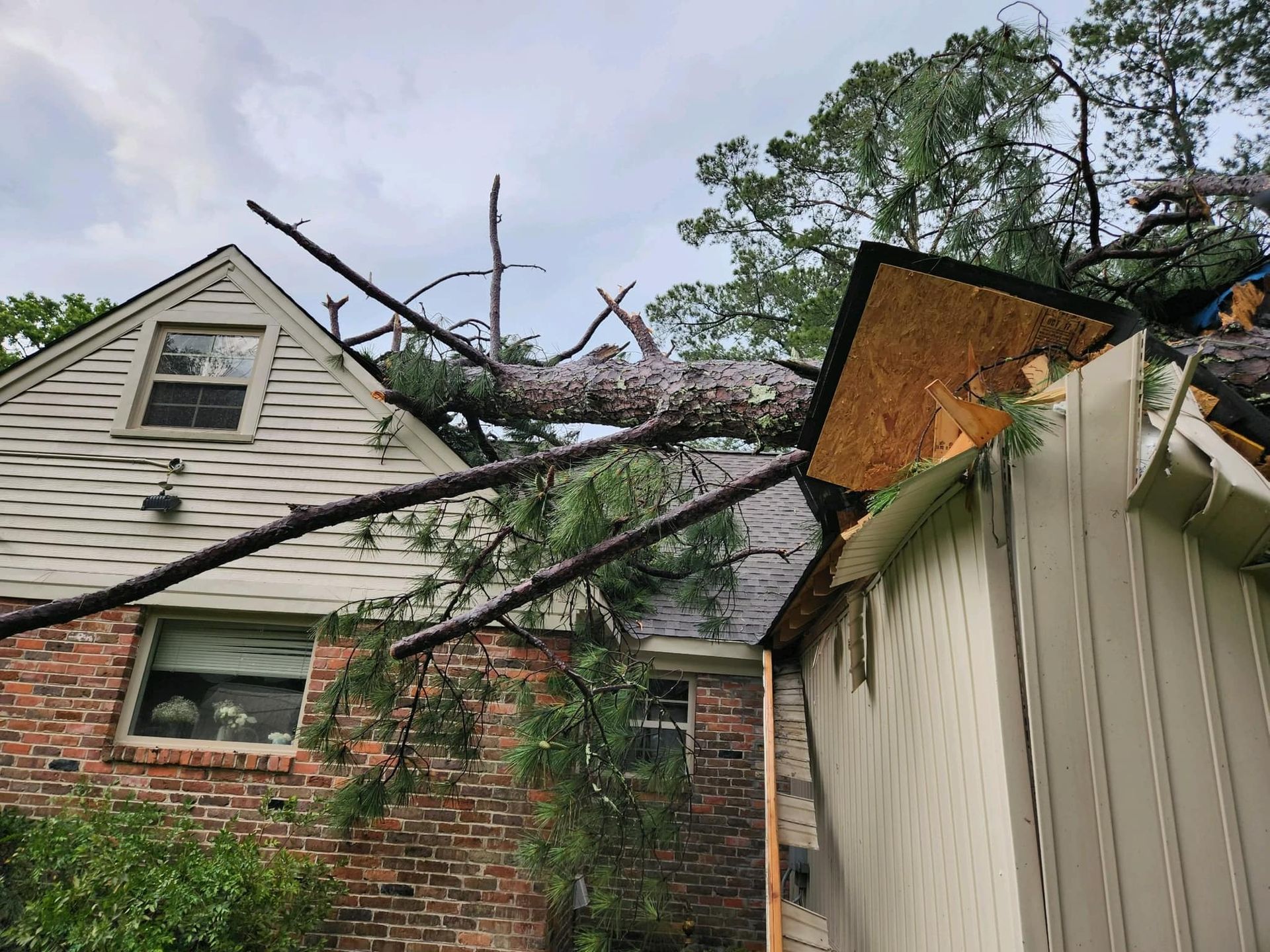 Tree branch fallen on house roof, causing damage. Beige siding, brick, cloudy sky.