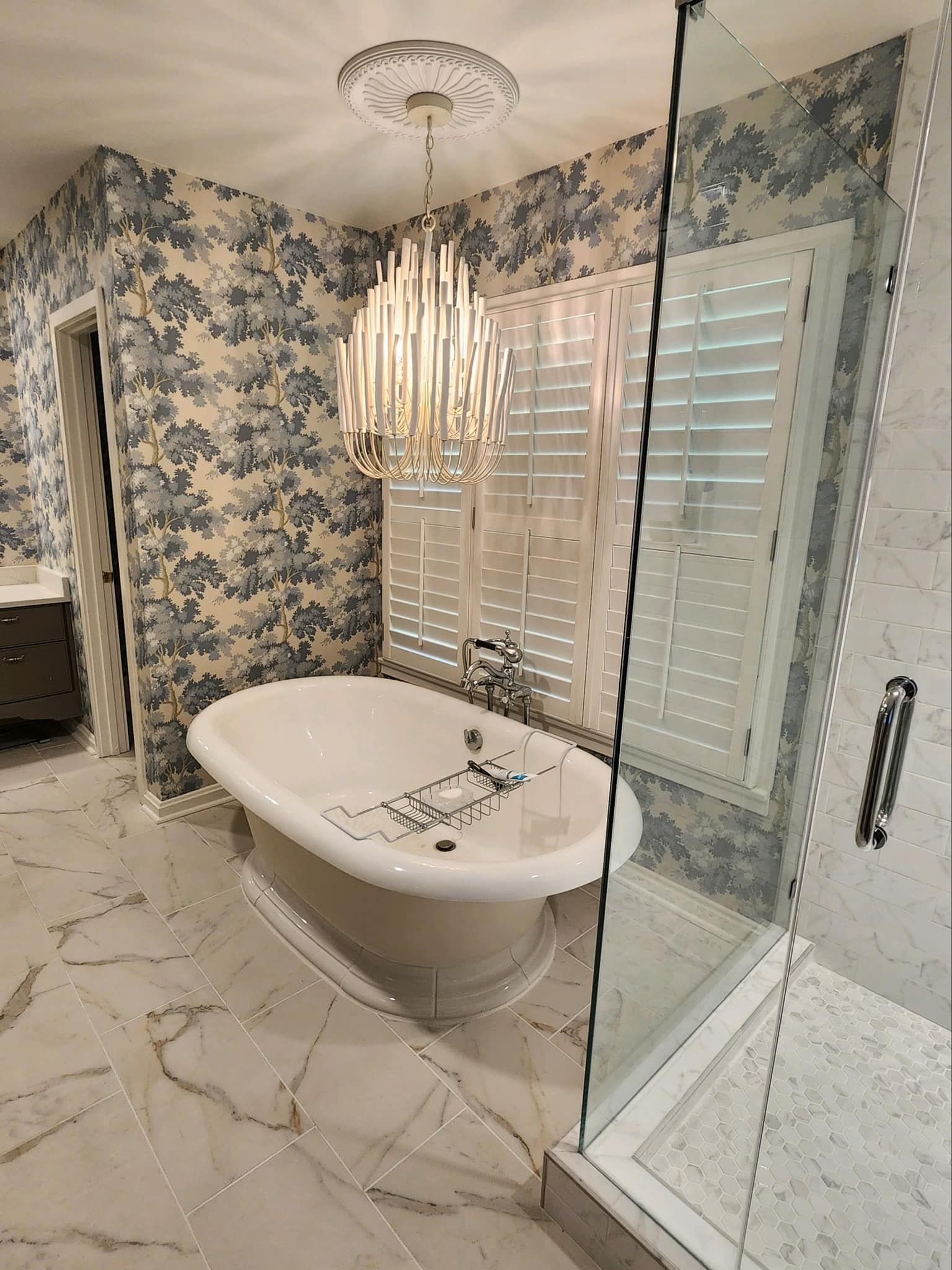 Luxurious bathroom with clawfoot tub, patterned wallpaper, marble floors, glass shower, and chandelier.