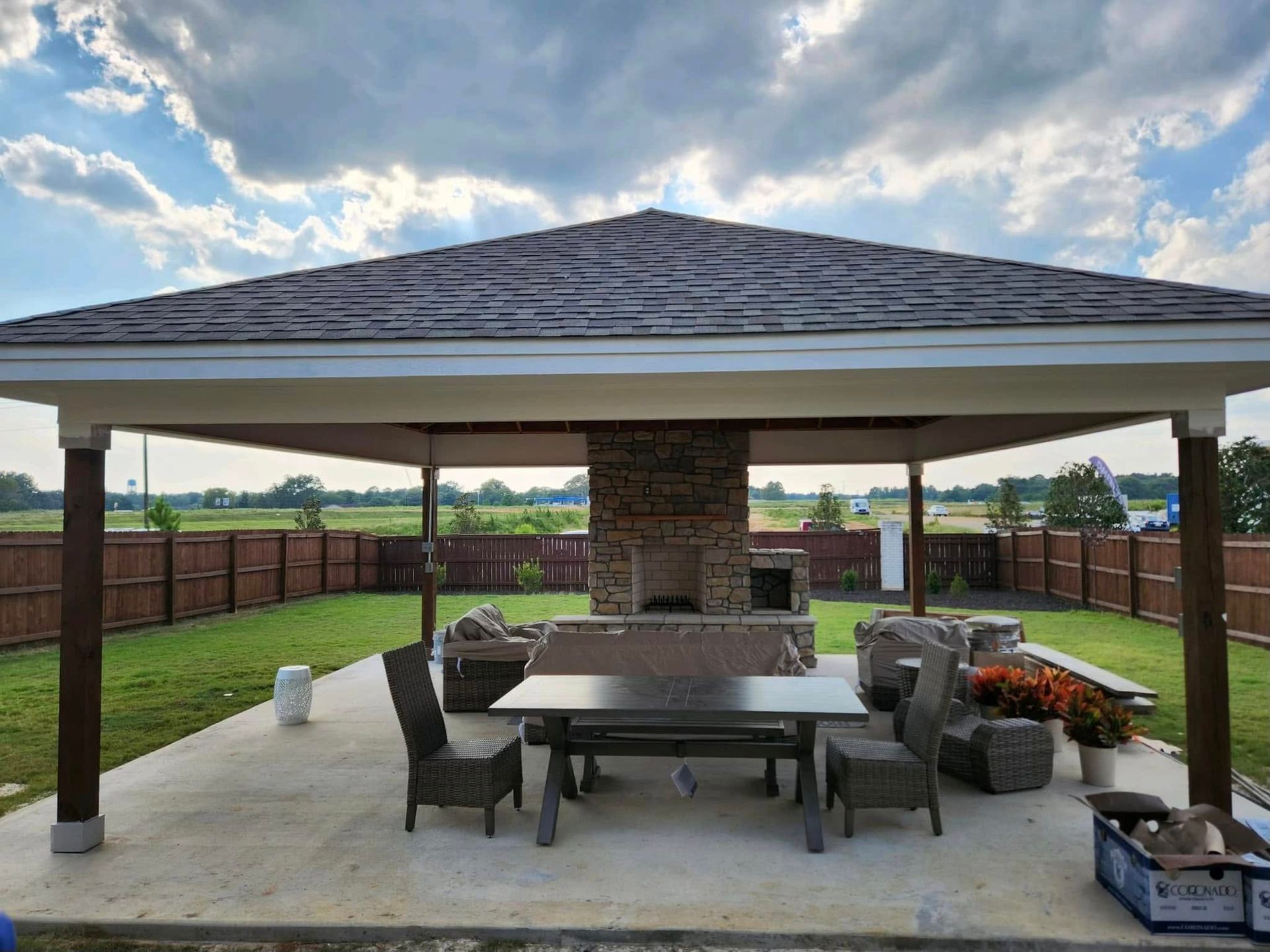 Outdoor pavilion with fireplace, seating, and table on a concrete patio, fenced backyard, cloudy sky.