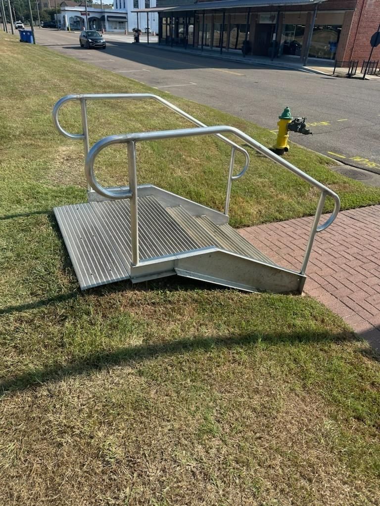 Metal ramp with handrails on grass, leading to brick sidewalk.