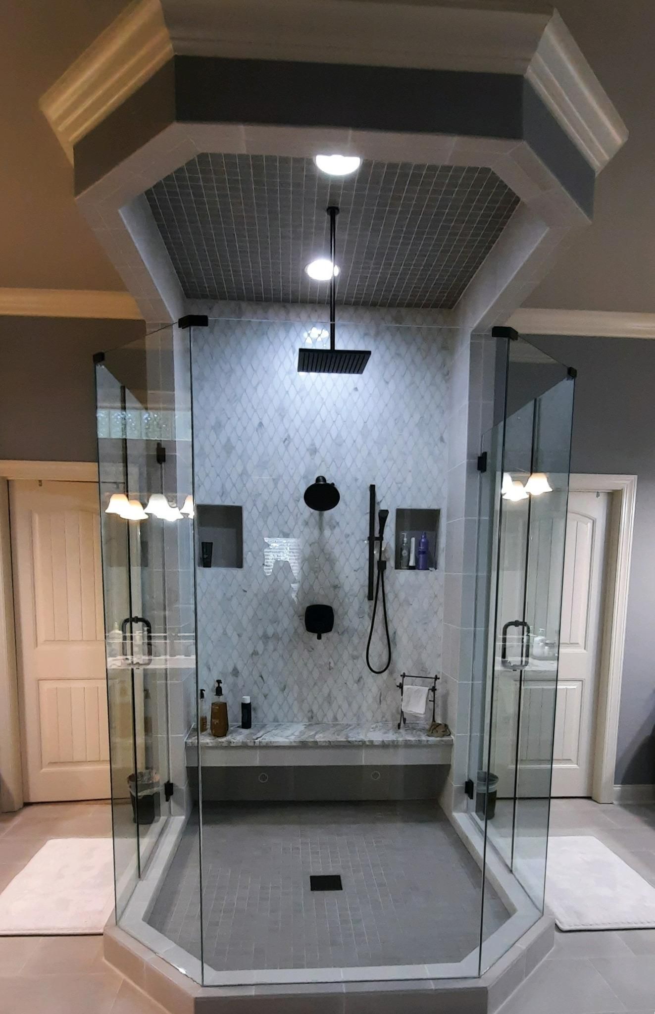 Spacious bathroom with glass shower enclosure, mosaic tile, and bench seating. Overhead lighting and gray tones.
