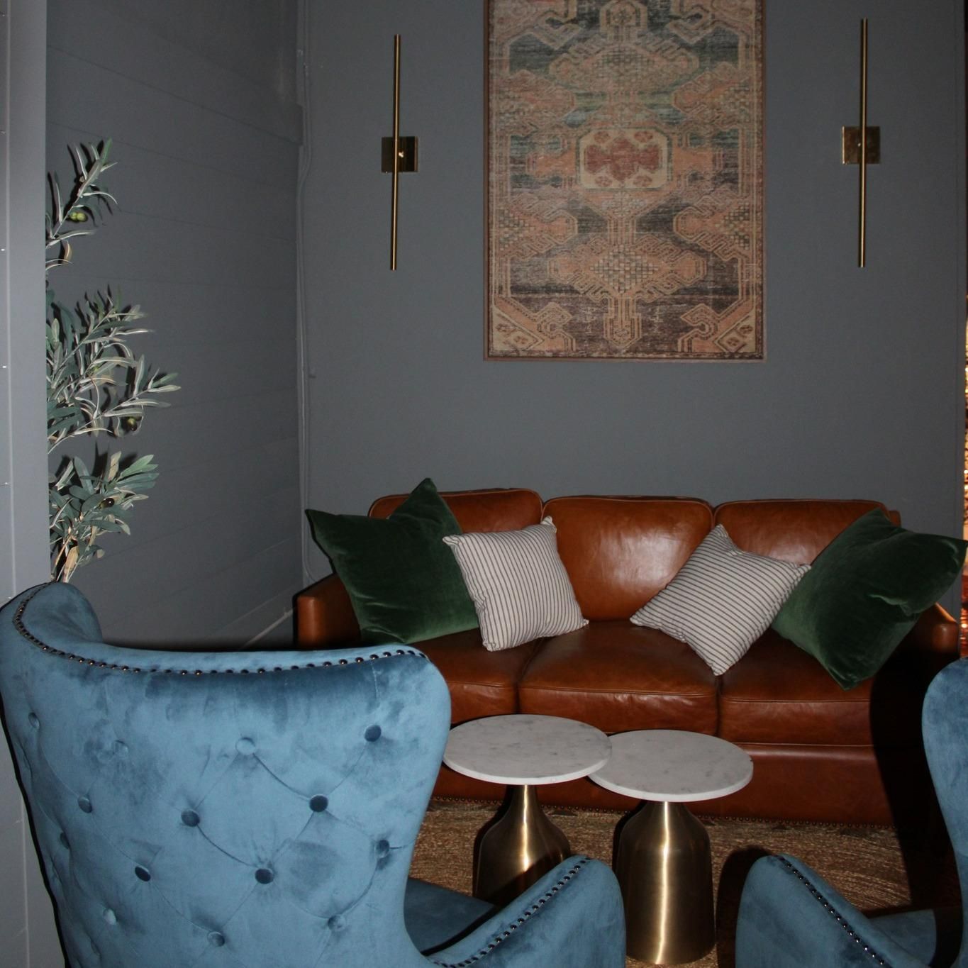 Cozy lounge area with a brown leather couch, teal chairs, and marble-topped tables. Dark blue walls, a rug, and wall art.