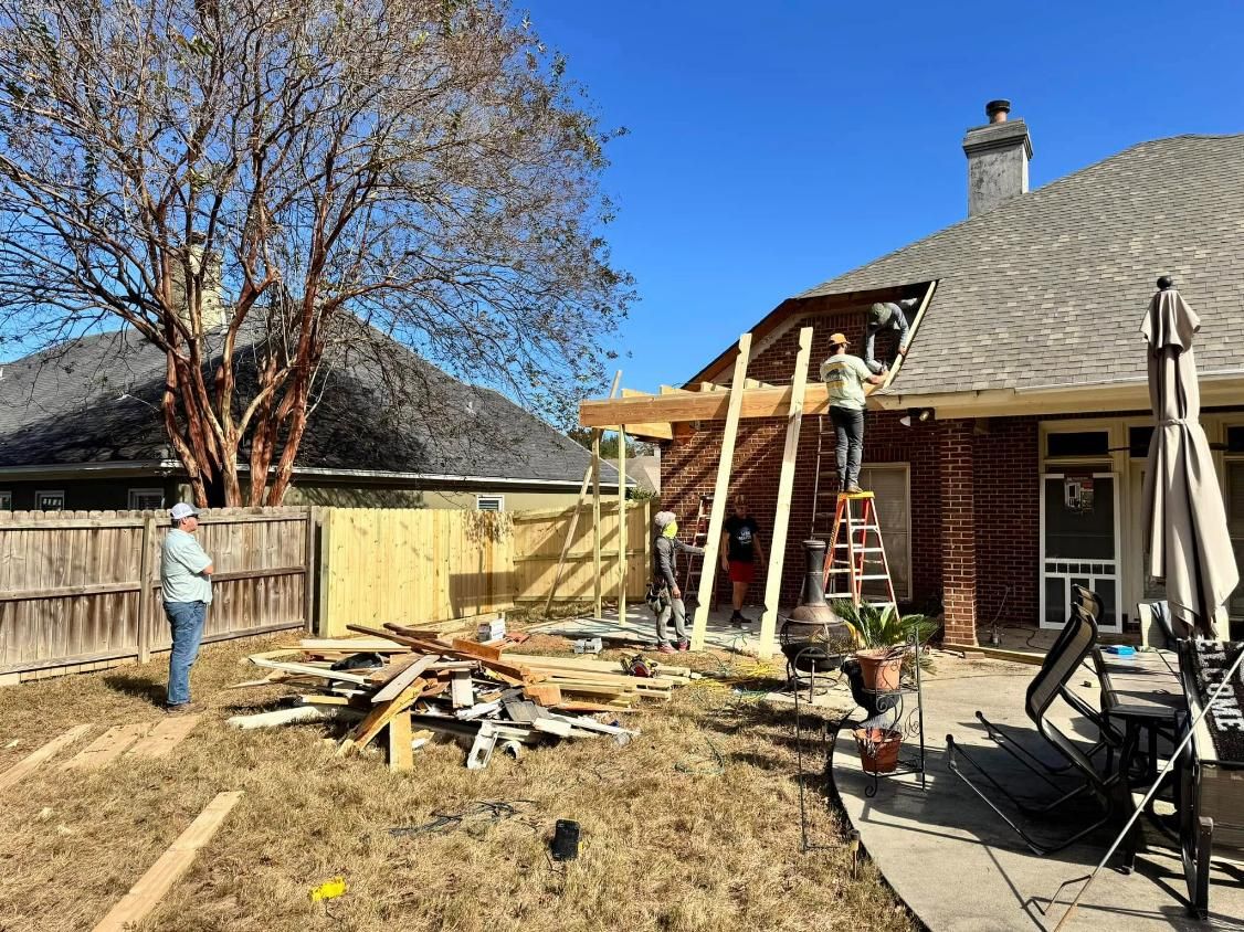Construction of a wooden pergola on a brick house patio. Several people work, with materials and tools strewn around.