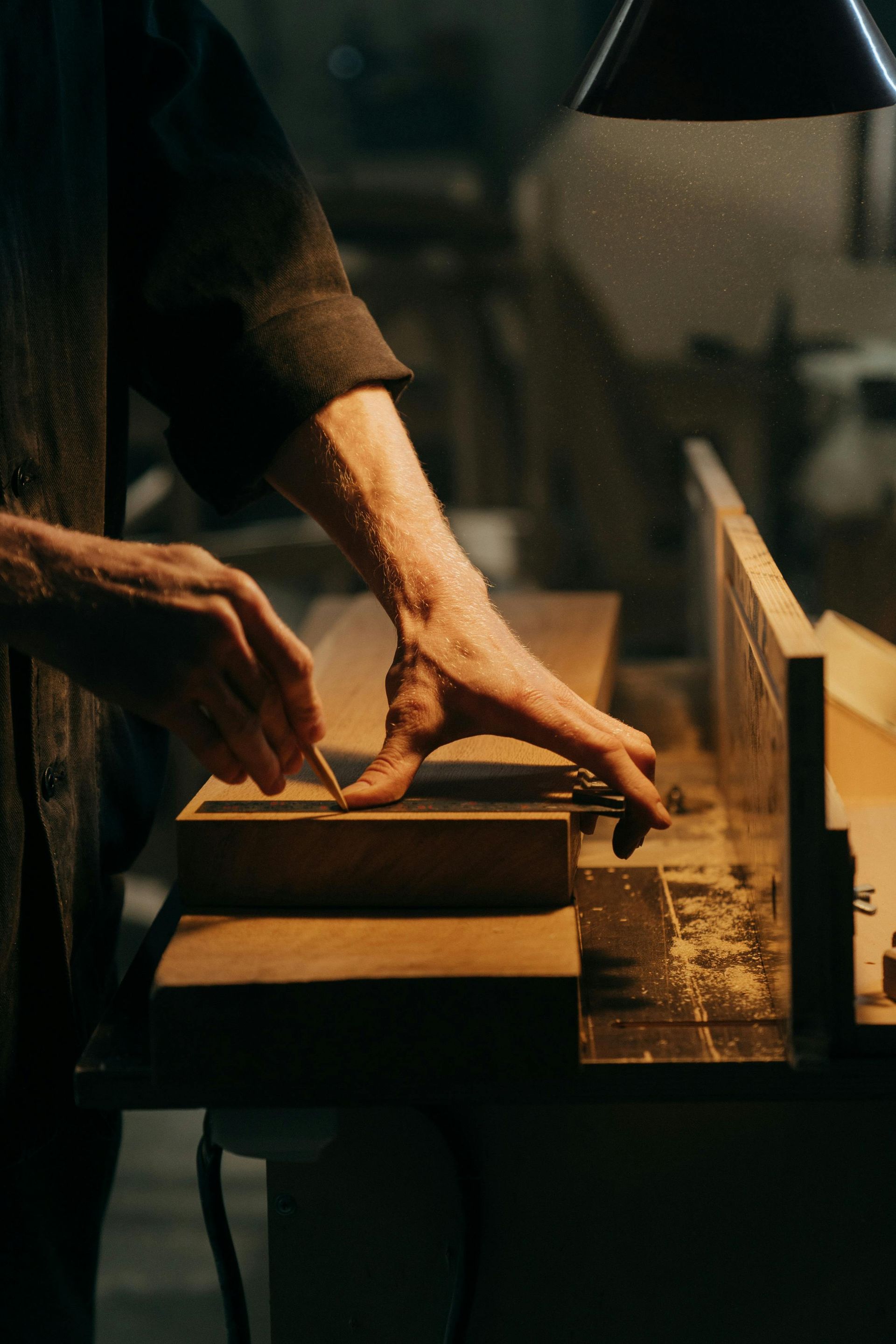 A person's hands guiding wood on a machine, illuminated by a lamp in a workshop.