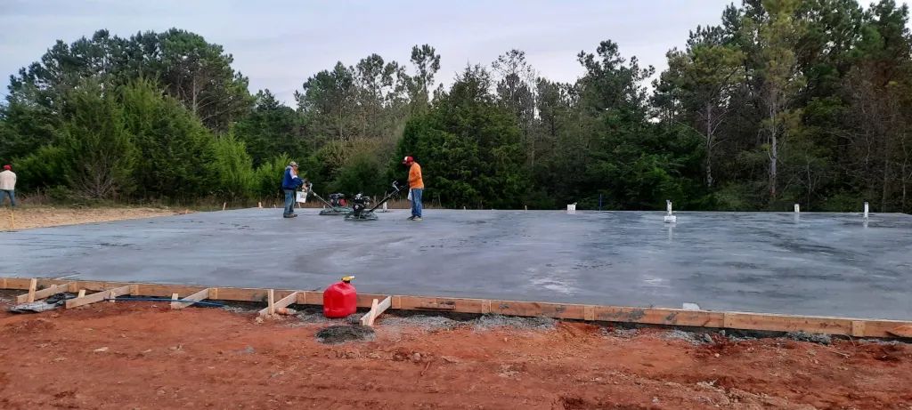 Workers on a poured concrete foundation, with trees in the background, a red container in the foreground.