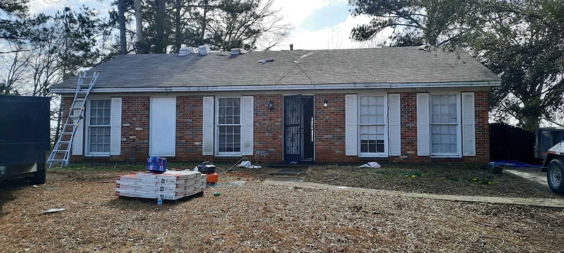 A brick house with a new roof, a ladder, and stacked shingles on the lawn.