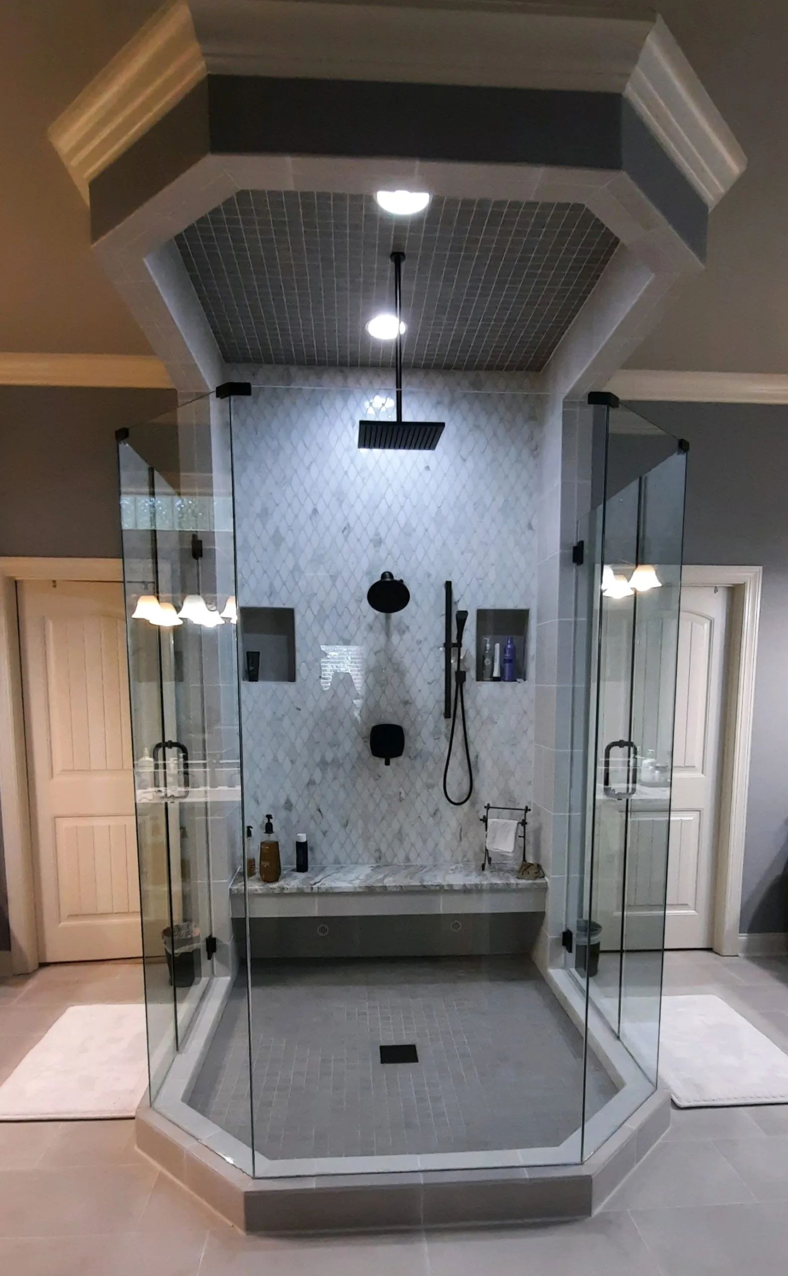 Luxurious, octagon-shaped shower with glass doors, built-in bench, and multiple showerheads. Gray tile, white accents, recessed lighting.