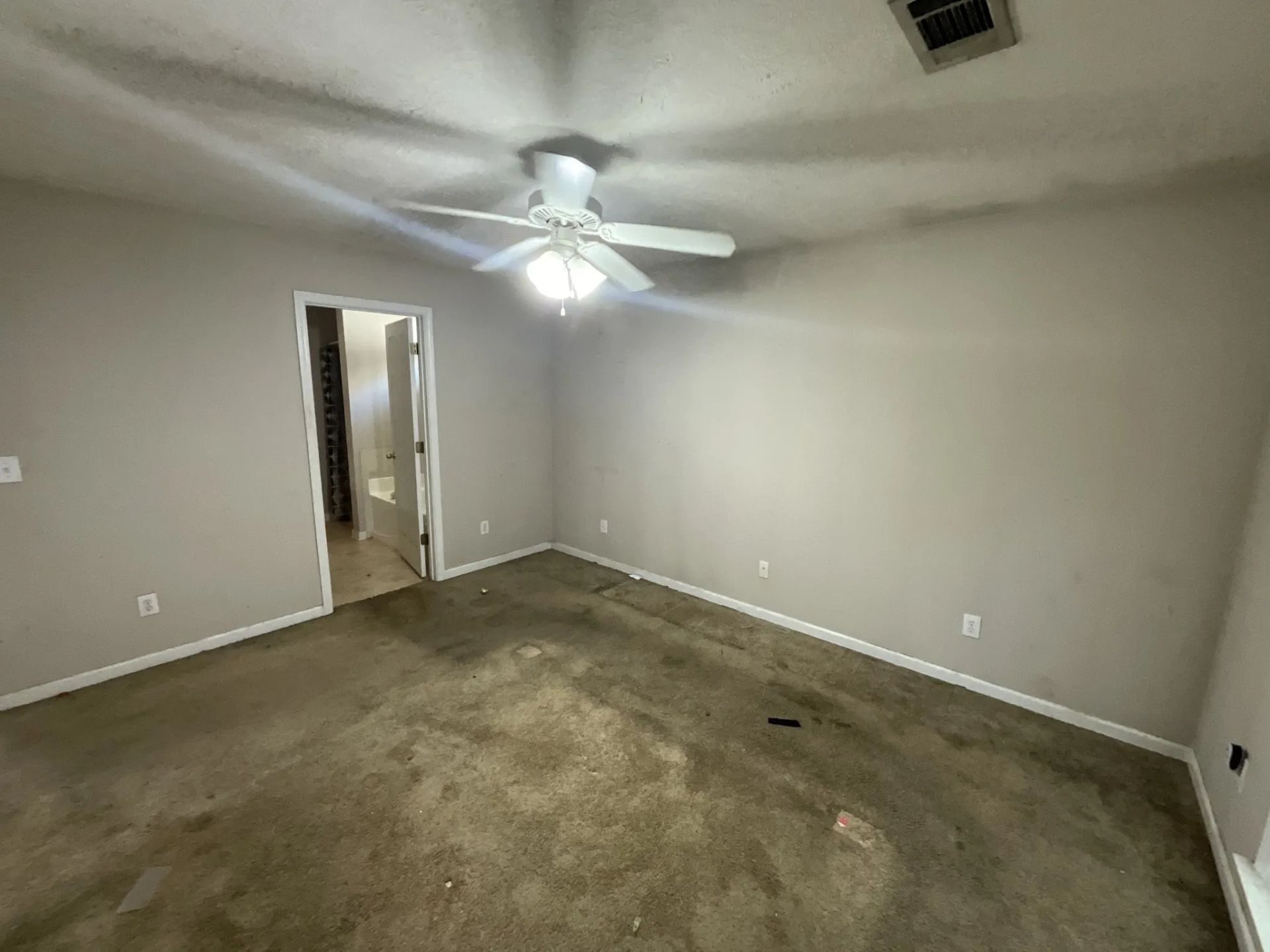 Empty bedroom with stained carpet, a ceiling fan, and a doorway to a bathroom.