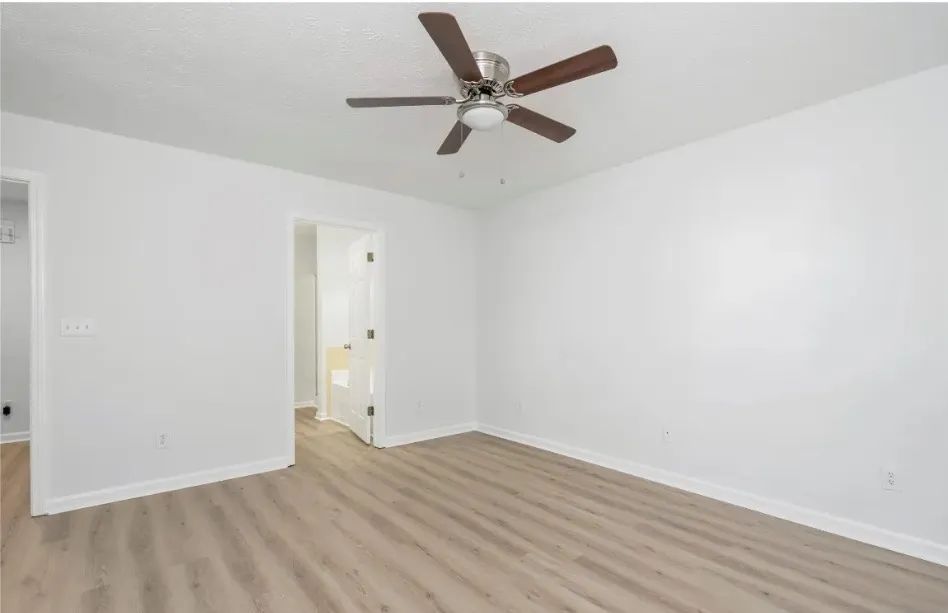Empty bedroom with light wood-look flooring, white walls, ceiling fan, and open doorway to another room.