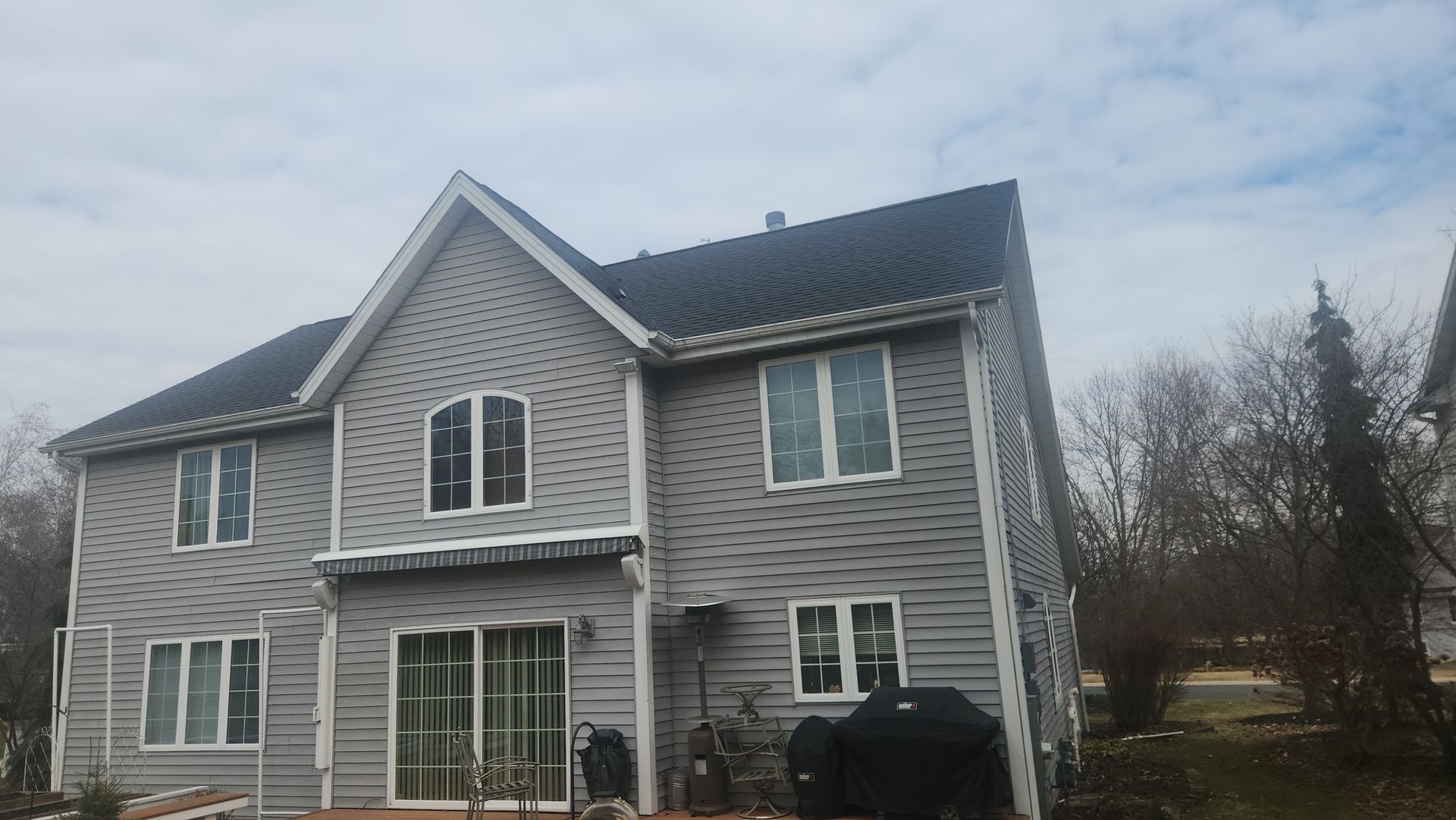 A large house with a black roof and grey siding has a grill in front of it.